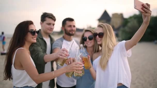 Grupp av vänner att ha kul njuter av en dryck och koppla av på stranden vid solnedgången i slow motion. Unga män och kvinnor dricka öl stående på en sand i den varma sommarkvällen. — Stockvideo