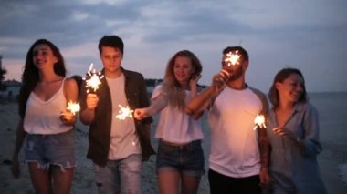 Arkadaşlar, yürüyüş bengal maytap ışıklar ellerinde ile deniz kenarında dans ve having fun gece boyunca parti. Havai fişek ile sahilde parti gençler. Ağır çekim steadycam vurdu.