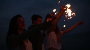 Arkadaşlar, yürüyüş bengal maytap ışıklar ellerinde ile deniz kenarında dans ve having fun gece boyunca parti. Havai fişek ile sahilde parti gençler. Ağır çekim steadycam vurdu.