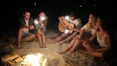 Gün batımı gün batımında kutluyor havai fişekleri ile plaj gör. Bengal ışıklar ve şarkı ile şenlik ateşi etrafında oturan kadın ve erkekler. Sakallı hippi gitar çalmak.
