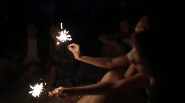Gün batımı gün batımında kutluyor havai fişekleri ile plaj gör. Bengal ışıklar ve şarkı ile şenlik ateşi etrafında oturan kadın ve erkekler. Sakallı hippi gitar çalmak.