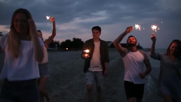 Amici che camminano, ballano e si divertono durante la festa notturna al mare con le luci scintillanti del bengala in mano. Giovani adolescenti che festeggiano sulla spiaggia con fuochi d'artificio. Scatto steadycam rallentato . — Video Stock