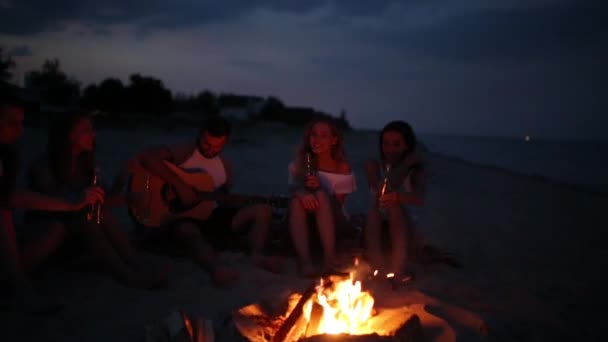 Beach party při západu slunce s ohněm. Přátelé, seděli kolem ohně, popíjeli pivo a zpívá na kytaru. Mladí muži a ženy drží skleněných lahví s nápoj zpěvné a fandění. — Stock video