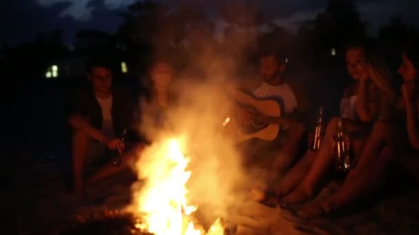 Strandfest ved solnedgang med bål. Venner sidder ved bålet, drikker øl og synger for guitaren. Mænd og kvinder holder glasflasker med drikkevare singalong, skægget fyr spiller guitar . – Stock-video