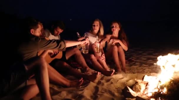 Fête à la plage au coucher du soleil avec feu de joie. Des amis assis autour du feu de joie, buvant de la bière et chantant à la guitare. Les jeunes hommes et les femmes tiennent des bouteilles en verre avec des boissons singalong et acclamant . — Video