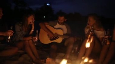 Şenlik ateşi ile gün batımında kumsal partisi. Şenlik ateşi oturan, bira içip şarkı gitar için arkadaşlar. Kadın ve erkek içecek singalong, sakallı adam gitar çalmak ile cam biberon.