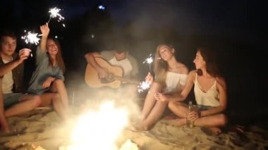Gün batımı gün batımında kutluyor havai fişekleri ile plaj gör. Bengal ışıklar ve şarkı ile şenlik ateşi etrafında oturan kadın ve erkekler. Sakallı hippi gitar çalmak.