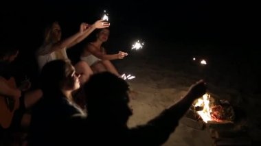 Gün batımı gün batımında kutluyor havai fişekleri ile plaj gör. Bengal ışıklar ve şarkı ile şenlik ateşi etrafında oturan kadın ve erkekler. Sakallı hippi gitar çalmak.