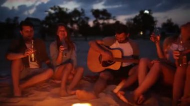 Şenlik ateşi ile gün batımında kumsal partisi. Şenlik ateşi oturan, bira içip şarkı gitar için arkadaşlar. Genç erkek ve kadınların içecek singalong ve tezahürat ile cam biberon.