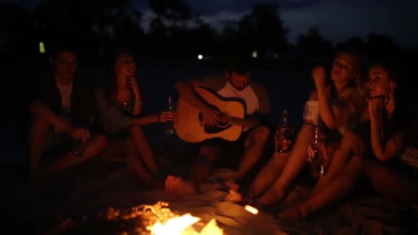 Fiesta en la playa al atardecer con hoguera. Amigos sentados alrededor de la hoguera, bebiendo cerveza y cantando a la guitarra. Hombres y mujeres sostienen botellas de vidrio con bebida singalong, barbudo chico tocando la guitarra . — Vídeos de Stock