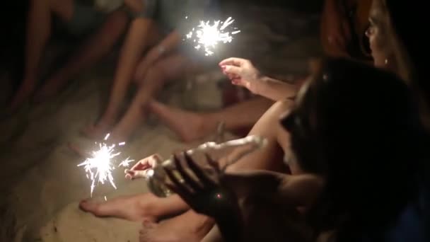 Друзья на пляже с бенгальскими огнями, празднующими закат. Молодые люди сидят у костра с бенгальскими огнями и поют. Бородатый хипстер играет на гитаре . — стоковое видео