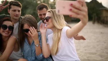Bir grup genç arkadaşıyla sahilde bir selfie alarak bir kadeh. Erkekler ve kadınlar sıcak yaz akşamı bir kum üzerine oturan fotoğraf çekmek.