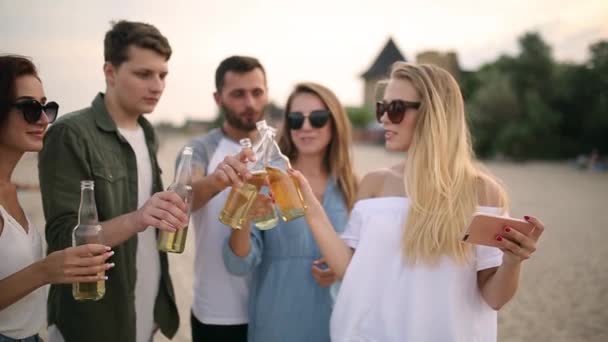 Grupo de amigos divirtiéndose disfrutando de una bebida y relajándose en la playa al atardecer en cámara lenta. Hombres y mujeres jóvenes beben cerveza de pie sobre una arena en la cálida noche de verano . — Vídeos de Stock