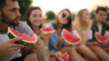 Mutlu arkadaş tatile Sandy Beach üzerinde oturan karpuz yeme. Kot şort giyen kadın ve erkekler. Dostluk ve yaz konsepti