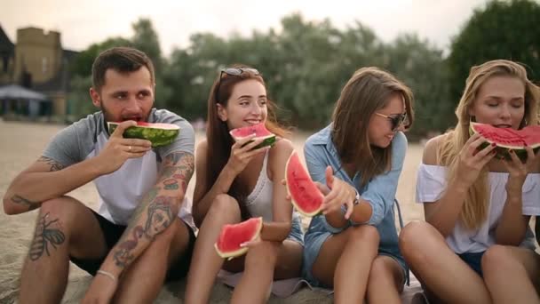 Щасливі друзів їсть кавун, сидячи на піщаний пляж у відпустці. Молоді чоловіки і жінки в шортах сині джинси. Дружба і літніх концепції — стокове відео
