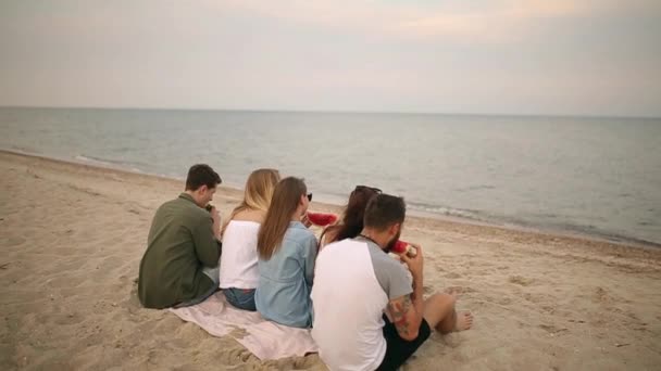 Happy Friends jedzenie arbuza, siedząc na piaszczystej plaży na wakacjach. Młodych kobiet i mężczyzn na sobie niebieskie dżinsy szorty. Przyjaźń i koncepcja lato — Wideo stockowe