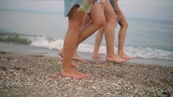 Ізольований вигляд трьох пар тонких жіночих ніг зі скляними пляшками напою в руках, що ступають на пляж біля моря під час заходу сонця в повільному русі. Молоді жінки п'ють пиво, що йде на пісок. Без обличчя . — стокове відео