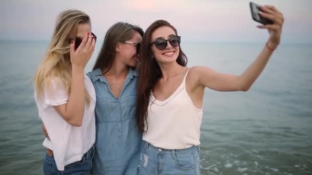 Tres mujeres jóvenes tomando un selfie en la playa con vistas al mar. Los amigos sonríen mirando a la cámara. Chicas con pantalones cortos de mezclilla azul y vestido. Concepto de ropa de algodón ecológico . — Vídeos de Stock