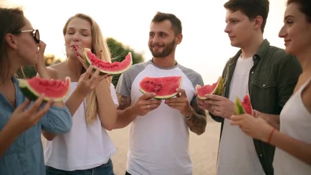 Счастливые друзья едят арбуз, стоя на песчаном пляже и болтая. Молодые мужчины и женщины в синих джинсовых шортах у моря в отпуске. Концепция дружбы и лета — стоковое видео