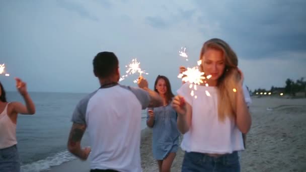 Freunde spazieren, tanzen und amüsieren sich bei einer nächtlichen Party am Meer mit Bengalischen Wunderkerzen in der Hand. Jugendliche feiern am Strand mit Feuerwerk. Schwebestativ in Zeitlupe geschossen. — Stockvideo