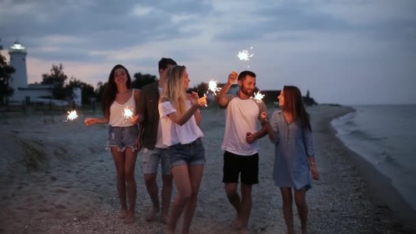 Amigos caminhando, dançando e se divertindo durante a festa à beira-mar com luzes brilhantes de bengala em suas mãos. Jovens adolescentes festejando na praia com fogos de artifício. câmera lenta steadycam tiro . — Vídeo de Stock