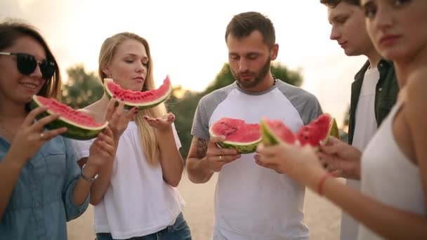 Felices amigos comiendo sandía de pie en Sandy Beach y charlando. Hombres y mujeres jóvenes con pantalones cortos vaqueros azules cerca del mar de vacaciones. Concepto de amistad y verano — Vídeos de Stock