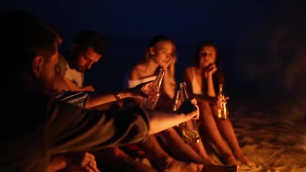 Festa de praia ao pôr do sol com fogueira. Amigos sentados ao redor da fogueira, bebendo cerveja e cantando para a guitarra. Jovens homens e mulheres seguram garrafas de vidro com bebida singalong e torcida . — Vídeo de Stock