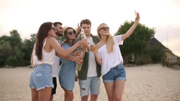 Skupina přátel baví užívat nápoj a odpočinek na pláži při západu slunce v pomalém pohybu. Mladí muži a ženy pijí pivo stojí na písku v teplé letní večery. — Stock video