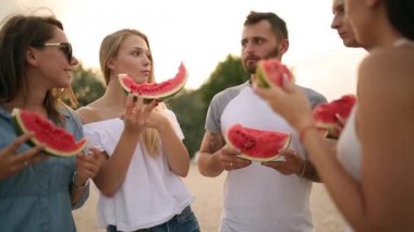 Mutlu arkadaş karpuz ayakta yemek Sandy Beach ve sohbet. Tatile denize yakın kot şort giyen kadın ve erkekler. Dostluk ve yaz konsepti
