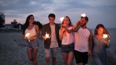 Arkadaşlar, yürüyüş bengal maytap ışıklar ellerinde ile deniz kenarında dans ve having fun gece boyunca parti. Havai fişek ile sahilde parti gençler. Ağır çekim steadycam vurdu.