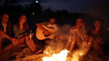 Şenlik ateşi ile gün batımında kumsal partisi. Şenlik ateşi oturan, bira içip şarkı gitar için arkadaşlar. Kadın ve erkek içecek singalong, sakallı adam gitar çalmak ile cam biberon.