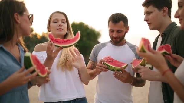 Happy Friends Eating Watermelon Em pé em Sandy Beach e conversando. Homens e mulheres jovens vestindo calções de calça azul perto do mar em férias. Amizade e Conceito de Verão — Vídeo de Stock
