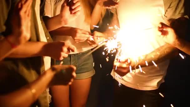 Amici in piedi intorno al fuoco più leggero durante la festa notturna al mare per accendere scintille bengala luci nelle loro mani. Giovani adolescenti che festeggiano sulla spiaggia con fuochi d'artificio. Scatto steadycam rallentato — Video Stock