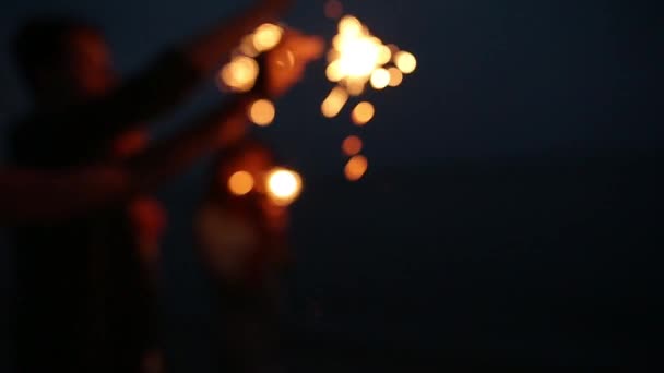 Tir au bokeh au ralenti. Amis marchant, dansant et s'amusant pendant la fête de nuit au bord de la mer avec des lumières scintillantes bengale dans leurs mains. Jeunes adolescents faisant la fête sur la plage avec des feux d'artifice . — Video