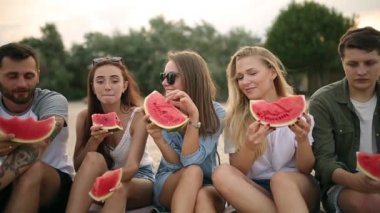 Mutlu arkadaş tatile Sandy Beach üzerinde oturan karpuz yeme. Kot şort giyen kadın ve erkekler. Dostluk ve yaz konsepti