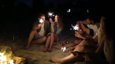 Gün batımı gün batımında kutluyor havai fişekleri ile plaj gör. Bengal ışıklar ve şarkı ile şenlik ateşi etrafında oturan kadın ve erkekler. Sakallı hippi gitar çalmak.