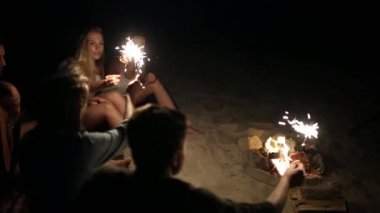 Gün batımı gün batımında kutluyor havai fişekleri ile plaj gör. Bengal ışıklar ve şarkı ile şenlik ateşi etrafında oturan kadın ve erkekler. Sakallı hippi gitar çalmak.