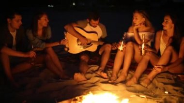 Şenlik ateşi ile gün batımında kumsal partisi. Şenlik ateşi oturan, bira içip şarkı gitar için arkadaşlar. Genç erkek ve kadınların içecek singalong ve tezahürat ile cam biberon.