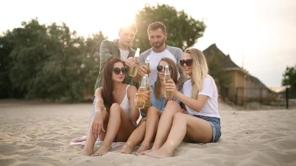 Group of friends having fun enjoying a beverage and relaxing on the beach at sunset in slow motion. Young men and women drink beer sitting on a sand in the warm summer evening. — Stock Video