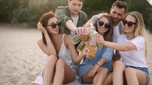 Group of friends having fun enjoying a beverage and relaxing on the beach at sunset in slow motion. Young men and women drink beer sitting on a sand in the warm summer evening. — Stock Video