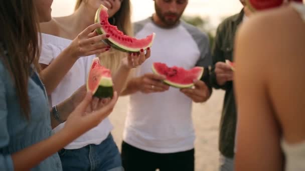 Boldog meg enni görögdinnye állandó homokos strand, és beszélgetni. Fiatal férfiak és nők visel kék farmer rövidnadrág-tenger közelében a nyaralás. Barátság és a nyári koncepció — Stock videók