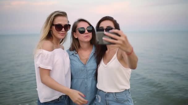 Три молодые женщины делают селфи на пляже с видом на море. Друзья улыбаются, глядя в камеру. Девушки в синих джинсовых шортах и платье. Концепция одежды из хлопка . — стоковое видео