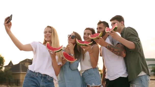 Przyjaciele jedzenie arbuza, stojąc na piaszczysta plaża i biorąc selfie z aparatem w telefonie. Młodych mężczyzn i kobiet na sobie niebieskie dżinsy szorty w pobliżu morza ans rozmowy. Zdrowe odżywianie, wegetarianką żywności — Wideo stockowe
