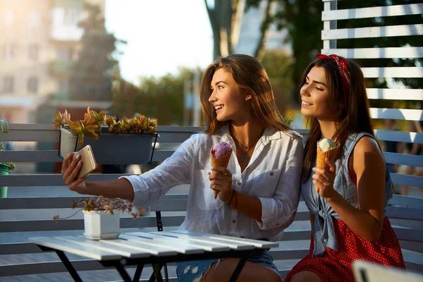 Portret van twee jonge vrouwen zitten samen eten ijs kegels en het nemen van de selfie foto op mobiele telefoon camera in straat zomerterras. Meisjes zich fotograferen met smartphone camera. — Stockfoto