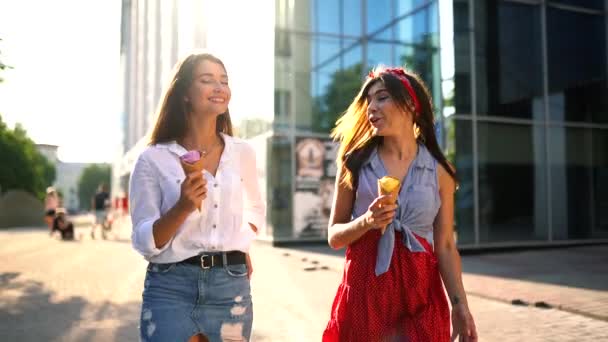 Dos amigas jóvenes divirtiéndose y comiendo helado. Alegre caucásico mujeres eati helado al aire libre caminando en la ciudad. Chicas bonitas posando con helado. Concepto horario de verano. Movimiento lento — Vídeos de Stock