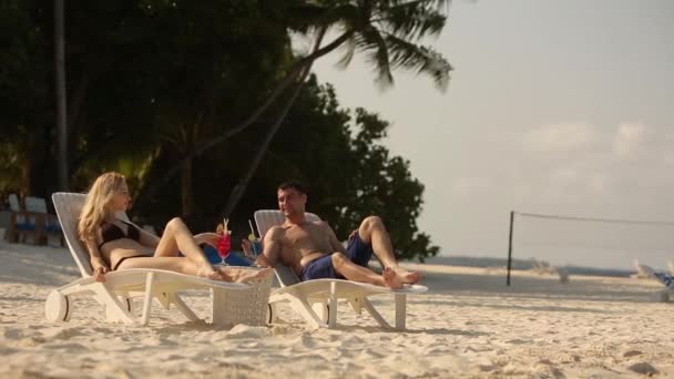 Молодая семейная пара загорает и пьет цветной коктейль на тропическом пляже. Любители медового месяца проводят экзотические каникулы, лежа на солнце на шезлонгах возле океана. — стоковое видео