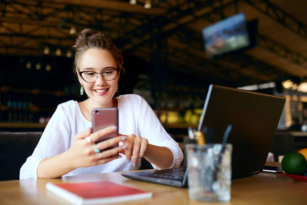 Mujer de negocios de raza mixta escribiendo un texto en el teléfono inteligente. Mujer asiática sosteniendo un teléfono celular moderno y un mensaje de escritura. Sonriente joven freelancer en gafas mirando el teléfono con portátil en la mesa . — Foto de Stock