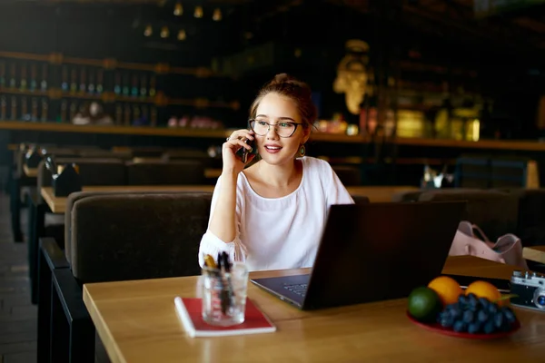 Joven freelancer trabajando con laptop hablando por celular con el cliente en la cafetería. Bastante asiática caucásica mixta mujer de negocios en gafas lleva a cabo negociaciones a través de llamada telefónica. Concepto multitarea . — Foto de Stock