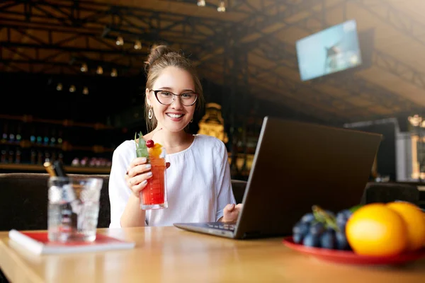 Smiling mixed race woman with cocktail in hand works with laptop. Businesswoman in glasses drinks juice for body hydration while working. Attractive designer quench thirst. Healthy lifestyle theme. — Stock Photo, Image
