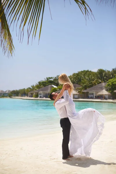 O noivo feliz mantém a noiva em suas mãos sob o arco da cerimônia de casamento na praia tropical da ilha nas Maldivas. Turquesa lagoa oceânica, areia branca e bangalôs de luxo resort spa no fundo. Lua de mel . — Fotografia de Stock